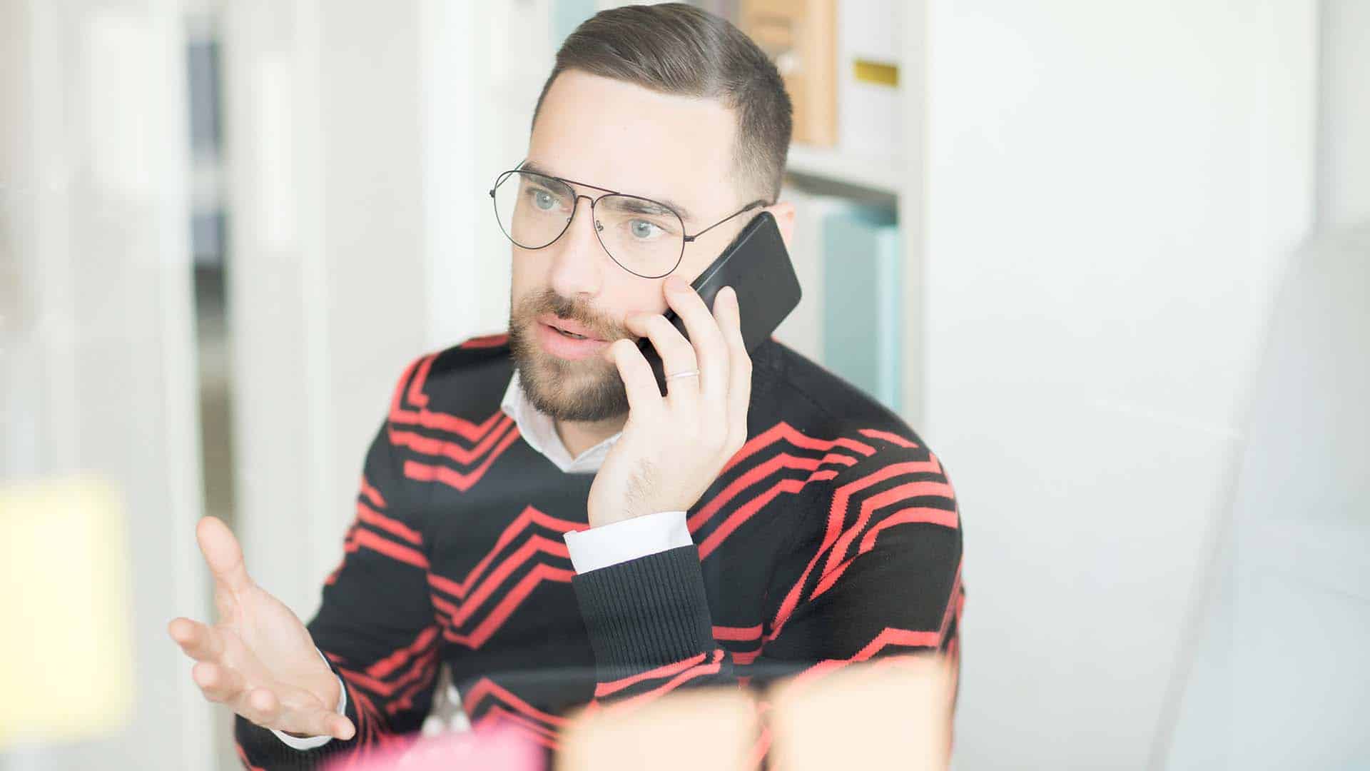 Confused outraged young male manager in glasses sitting in office of marketing company and talking by phone solving urgent issues