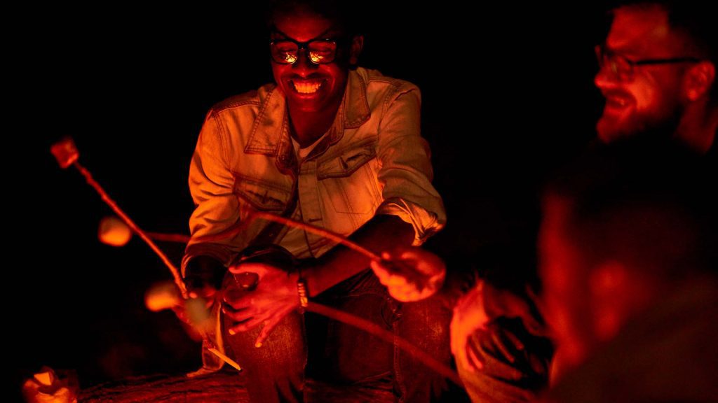 Cheerful happy young African man in eyeglasses roasting marshmallow while talking to close friends during night camping