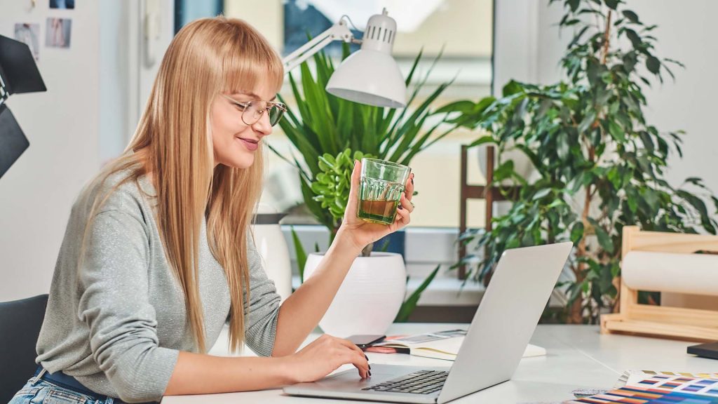 Happy smiling woman is holding a glass with drink while working on a laptop.