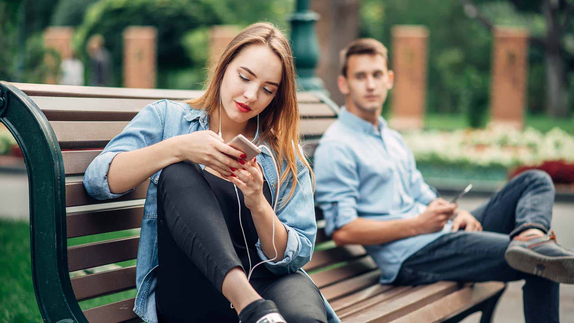 Phone addiction, smiling couple on the bench in park. Man and woman using their smartphones, social addicted people