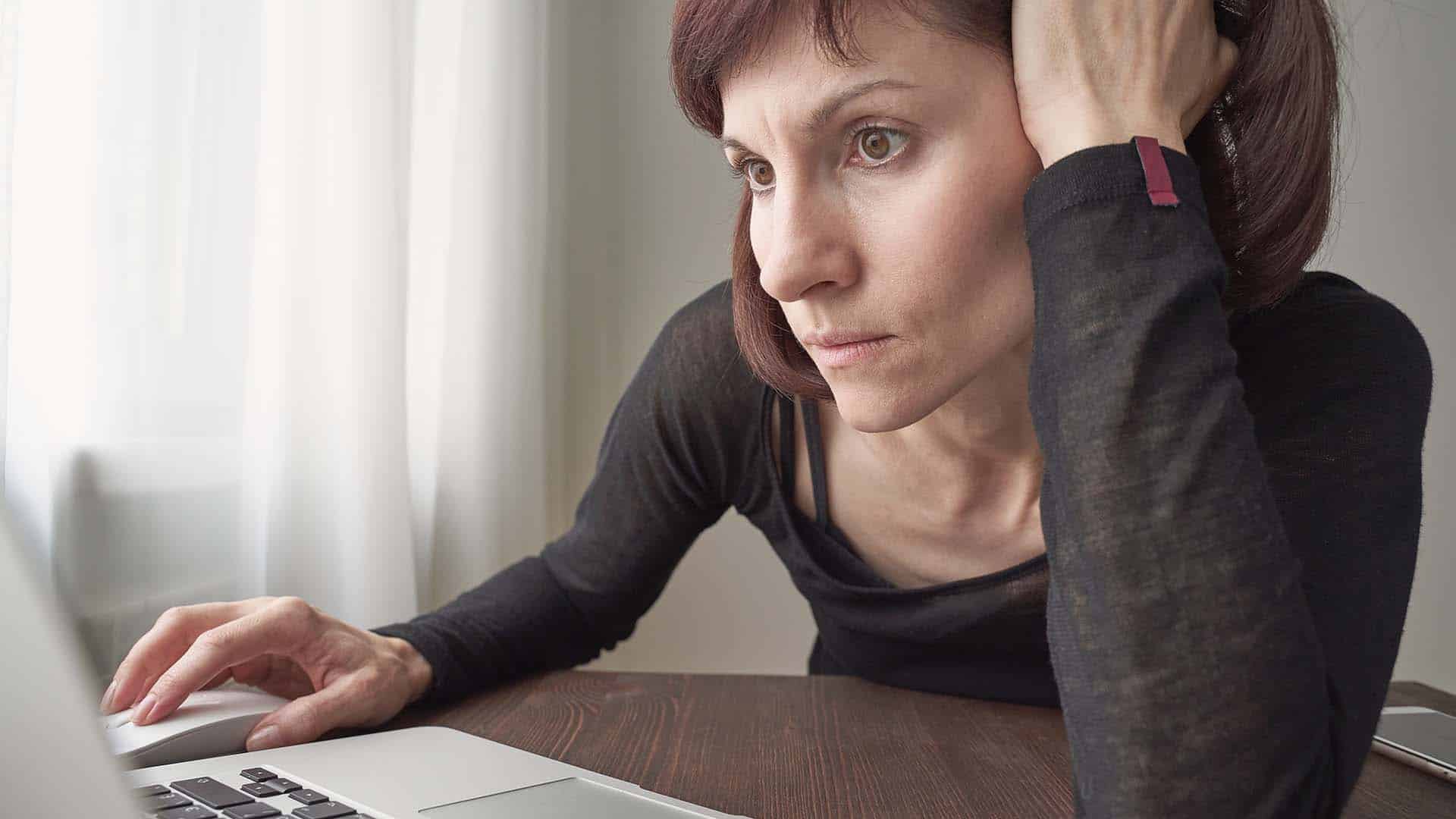 Woman is looking for information on Internet, nervous, focused. Concept for digital addiction. Adult mature female looks attentively and excitedly at computer. Anxiety, stress, fear.