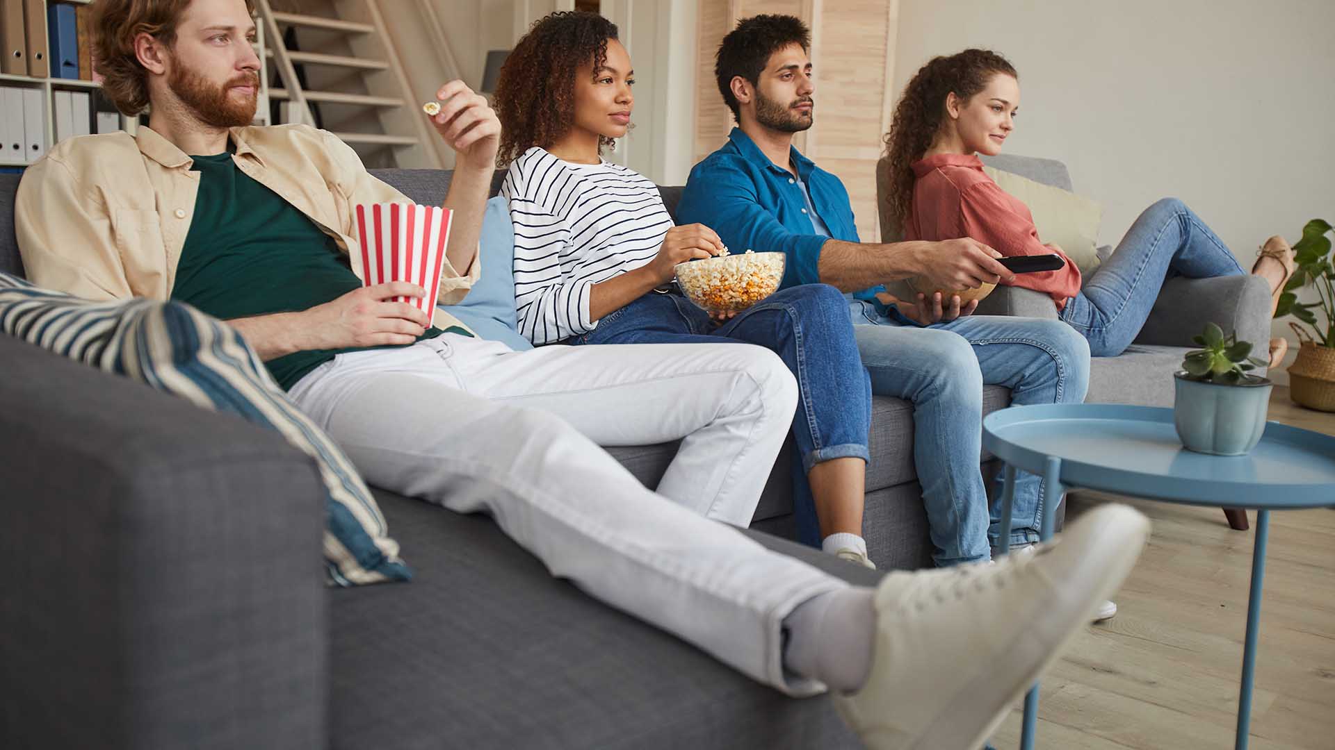 Low angle view at multi-ethnic group of friends watching TV together while sitting on comfortable sofa at home and enjoying snacks, copy space
