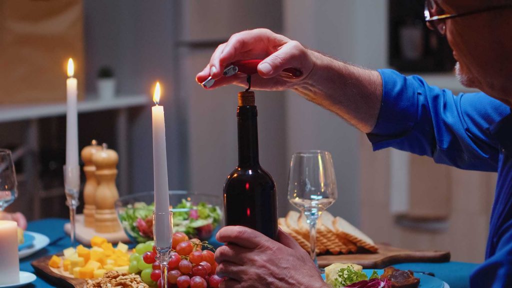 Close up of opening a bottle of wine during romantic dinner. Elderly mature old couple sitting at the table in kitchen, talking, enjoying the meal, celebrating their anniversary in the dining room.