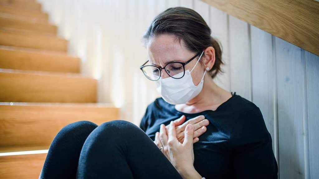 Sad and worried woman with face mask and chest pain sitting indoors at home, Corona virus and quarantine concept.