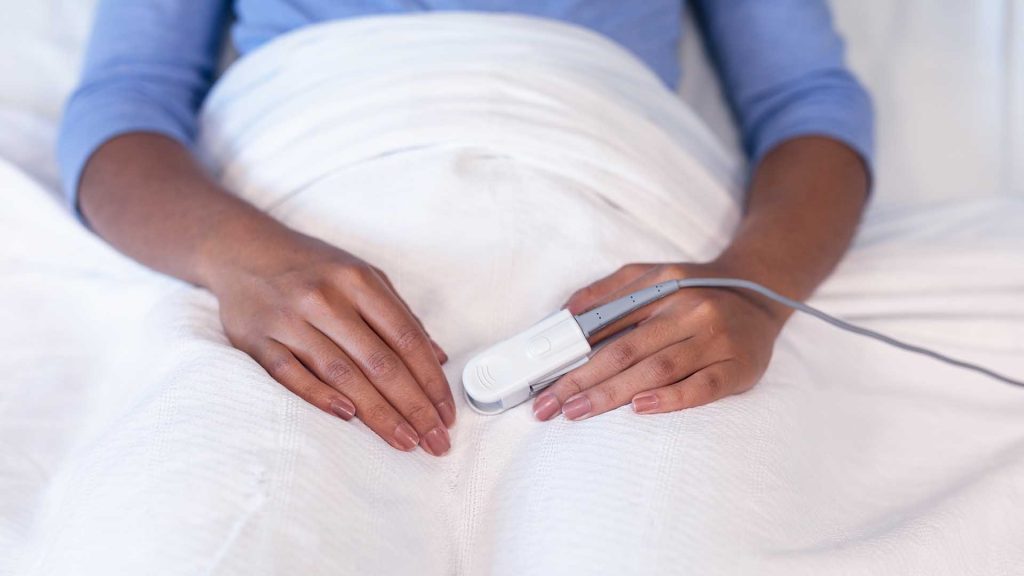 Midsection of mixed race female patient lying in hospital bed wearing fingertip pulse oximeter. medicine, health and healthcare services.