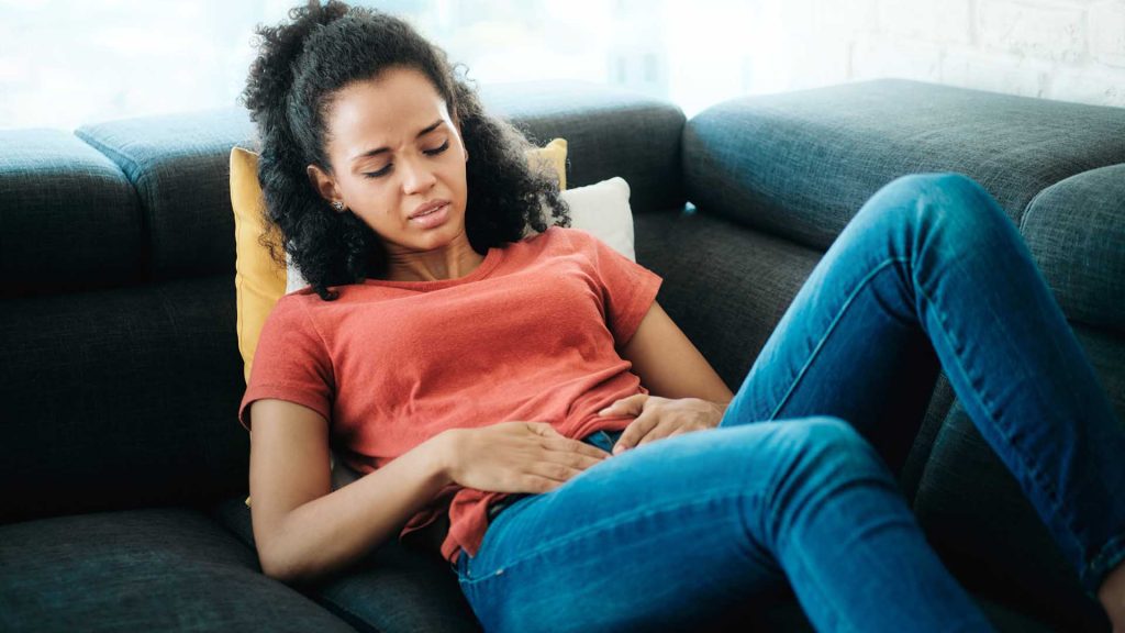Sad and sick young latina woman with menstrual pain. Ailing black girl having cramps during period and lying on sofa at home