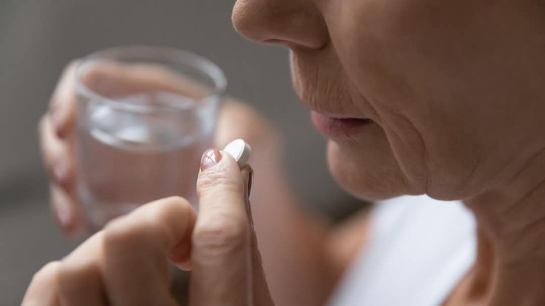 An older woman holds a pill and a glass of water to her mouth.