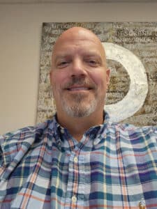 A selfie of Whit smiling at the camera, he has a shaved head with a goatee, and wearing a plaid shirt.